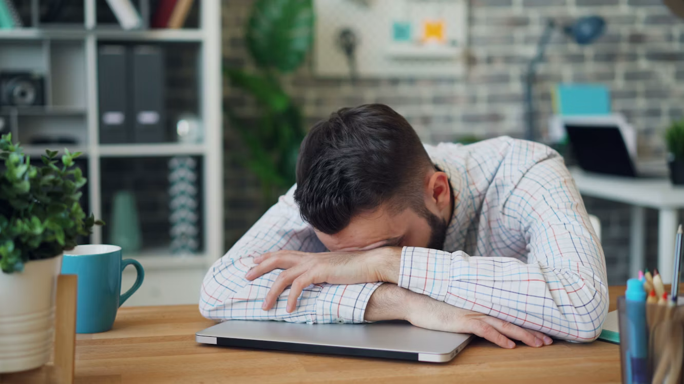 Homem exausto, com a cabeça apoiada nos braços sobre um notebook fechado, sentado à mesa em um escritório. Ao lado, há uma caneca azul, uma planta e materiais de trabalho, representando cansaço, estresse e sobrecarga profissional.
